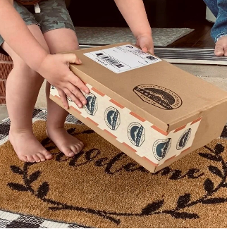 Two children picking up a Base Camp Treats Freeze Dried Subscription Box on a doormat with a brand logo.